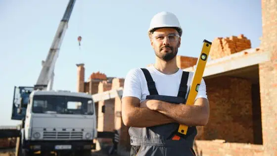 Fully licensed construction worker on site with a spirit level, construction crane, and brick building in the background, representing professional building and construction services.