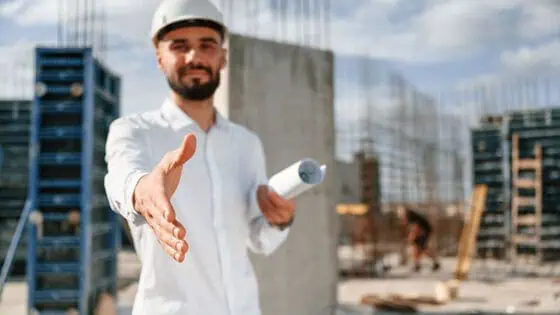 A construction project manager at a building site holding blueprints and extending hand for a handshake, with a background of modern skyscrapers under construction.
