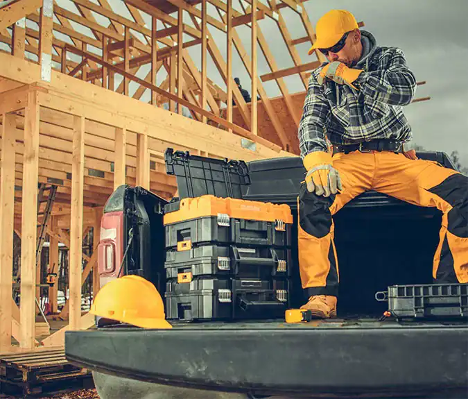 High-angle view of a construction worker sitting on the back of a pickup truck with tools and equipment, working on framing a wooden house under construction.