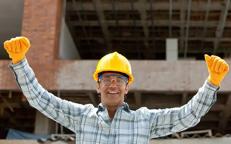 Happy construction worker wearing a yellow safety helmet and gloves, celebrating success at a construction site with a partially built structure in the background.