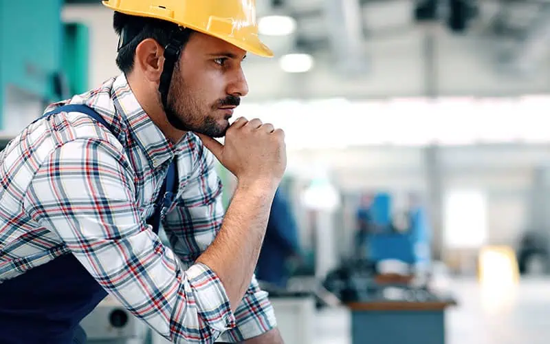 An industrial construction worker wearing a yellow safety helmet reviews blueprints in a manufacturing facility, emphasizing safety and planning for upcoming projects.