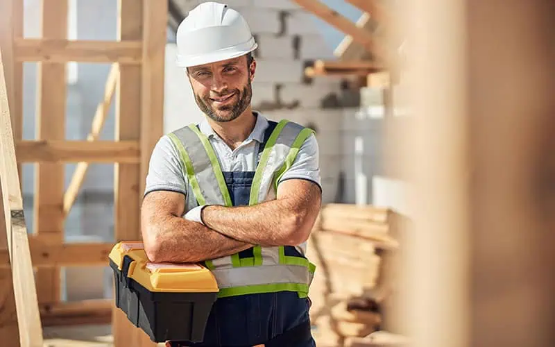 Construction worker wearing a white helmet and safety vest at a building site, smiling confidently with arms crossed, surrounded by wooden structures and construction materials.